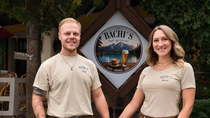 Ein Mann und eine Frau in Lederhosen und T-Shart halten sich an der Hand. Zwischen ihnen das Logo des Lokals