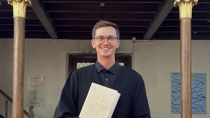 Ein junger Pfarrer steht zwischen Kirchenbänken