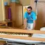 Ein junger, dunkelhaariger Mann trägt Kleidung für die Pflege, bezieht ein Bett und lacht in die Kamera