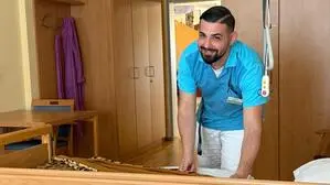 Ein junger, dunkelhaariger Mann trägt Kleidung für die Pflege, bezieht ein Bett und lacht in die Kamera