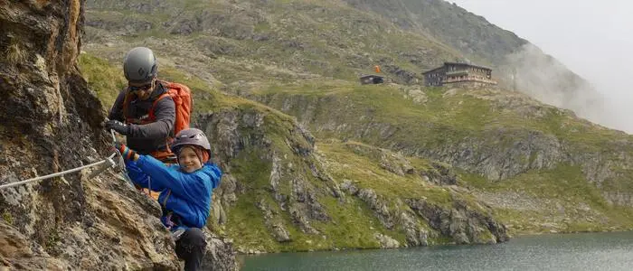 Ein Kind klettert auf einem Klettersteig. Hinter dem Kind ist ein Betreuer