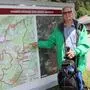 Ein Pensionist mit weißem Schnauzbart und Wanderkleidung steht im Wald und zeigt auf eine große Landkarte