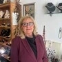 Eine Dame mit schulterlangen blonden Haaren und einer Brille sowie einer dunkelroten Jacke steht in ihrem Geschäft