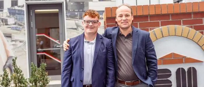Zwei Männer stehen neben einer Backsteinwand und lächeln für die Kamera