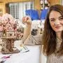 Auf einem Tisch steht ein künstlerisches Baumhaus, daneben ist eine braunhaarige Frau mit einer Pinzette und lächelt in die Kamera