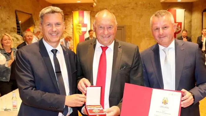 Drei Politiker in Anzügen halten eine Urkunde und eine Medaille in die Kamera.