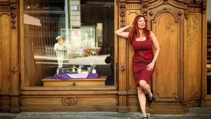 Sie trägt ein rotes, figurbetontes Kleid aus Trachtenstoff und posiert an die Wand gelehnt vor dem historischen Holzportal. Ihr Blick ist nach rechts gewandt