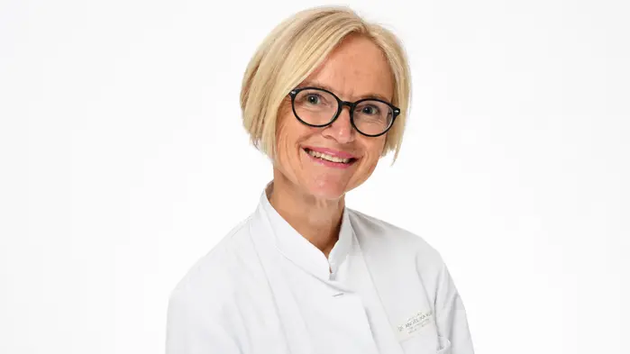 Eine blonde Frau mit Brille in einem Arztkittel lächelt in die Kamera