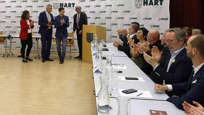 Das Bild zeigt die konstituierende Sitzung des Gemeinderats. Rechts sitzen einige Gemeinderäte an einem langen Tisch. Dahinter stehen andere vor einer Fotowand mit Hart-Logo
