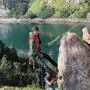 Eine Frau am Kletterseil in felsigem Gelände, dahinter Wald und ein See