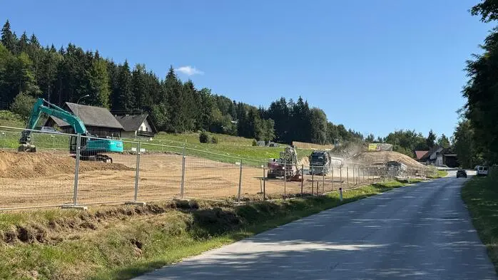 Eine eingezäunte Baustelle an einer Straße, im Hintergrund sieht man viele Bäume