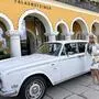 Eine blonde Frau mit Sonnenbrille und weißem Kostüm steht vor einem weißen Rolls Royce und lächelt in die Kamera