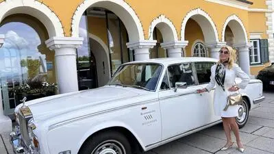 Eine blonde Frau mit Sonnenbrille und weißem Kostüm steht vor einem weißen Rolls Royce und lächelt in die Kamera