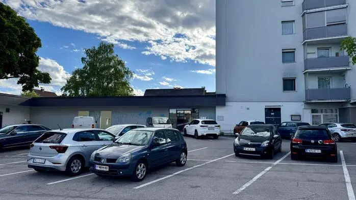 Blick auf einen Parkplatz auf dem mehrere Autos stehen, dahinter sieht man ein Gebäude, das verlassen wirkt, daneben erhebt sich ein mehrgeschossiges Wohnhaus
