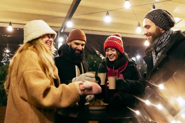 Cozy evening. Young cheerful men and women spending time together at winter traditional fair with hot drinks. Enjoying themselves, dressing warmly, looking at each other and laughing