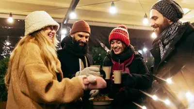Cozy evening. Young cheerful men and women spending time together at winter traditional fair with hot drinks. Enjoying themselves, dressing warmly, looking at each other and laughing