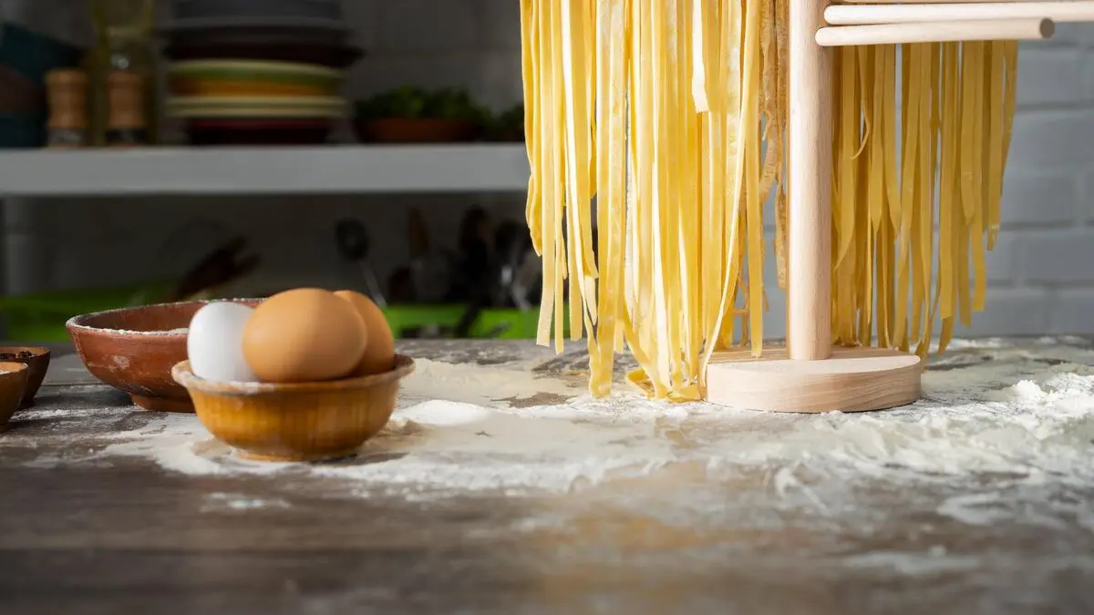 Tagliatelle hängen auf einer Holzvorrichtung, links daneben steht eine Schale mit Eiern