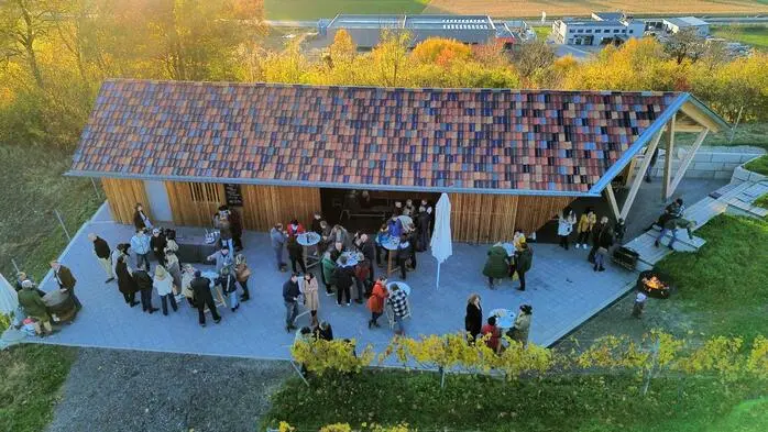 Aus der Vogelperspektive: Eine Location in Holzbauweise in einem Weingut bei schönem Wetter. Vor der Location stehen Tische und einige Menschen unterhalten sich.