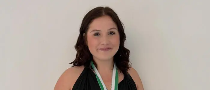 Eine junge Frau mit schwarzen Haaren hält eine Urkunde in die Kamera und hat eine Medaille um den Hals hängen