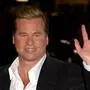 Das Bild zeigt Val Kilmer mit blonden, nach hinten gestylten Haaren, der lächelt und mit der rechten Hand winkt. Er trägt ein schwarzes Jackett über einem weißen Hemd. Der Hintergrund ist unscharf und wirkt dunkel mit einigen Lichtreflexen. | Kilmer wurde bekannt durch Rollen wie Jim Morrison in „The Doors“ und Batman.
