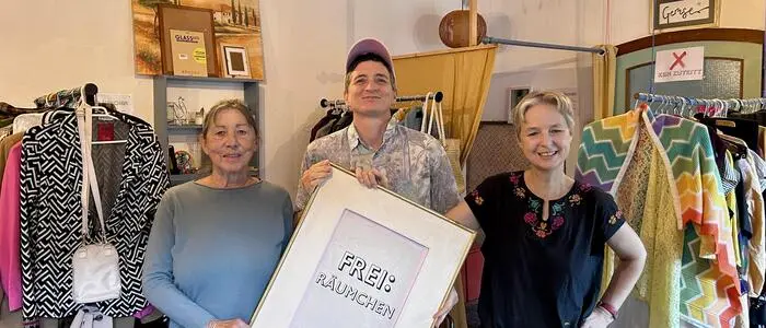 Drei Personen stehen in einem Laden mit Kleidung und halten ein Schild mit dem Text „Frei:räumchen“ hoch
