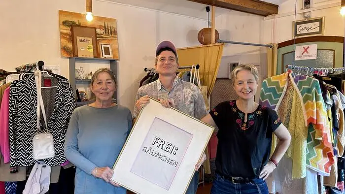 Drei Personen stehen in einem Laden mit Kleidung und halten ein Schild mit dem Text „Frei:räumchen“ hoch