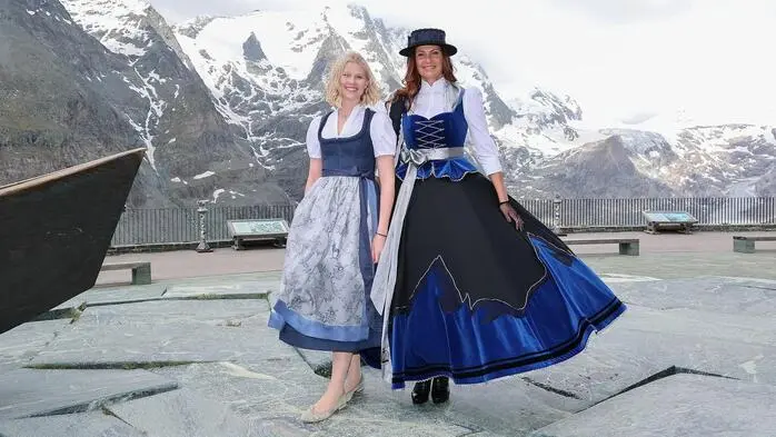 Zwei Frauen im blau-weißen Dirndl, dahinter eine Berglandschaft.