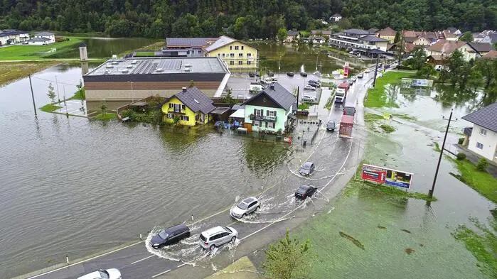 Luftbild zeigt überschwemmte Wiesen und Häuser sowie eine Straße, auf der sich Autos den Weg durchs Wasser bahnen