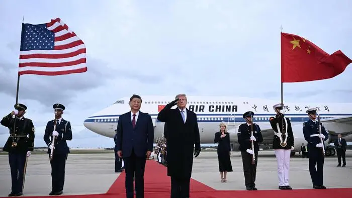 Donald Trump salutiert auf einem roten Teppich vor der chinesischen Präsidentenmaschine, rechts von ihm steht Xi Jinping. Weitere Personen stehen hinter bzw. neben ihnen.