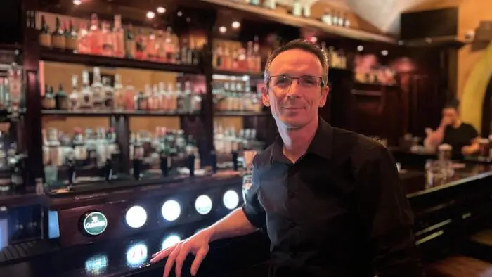 Ein Mann im schwarzen Hemd und mit Brille vor einer Bar