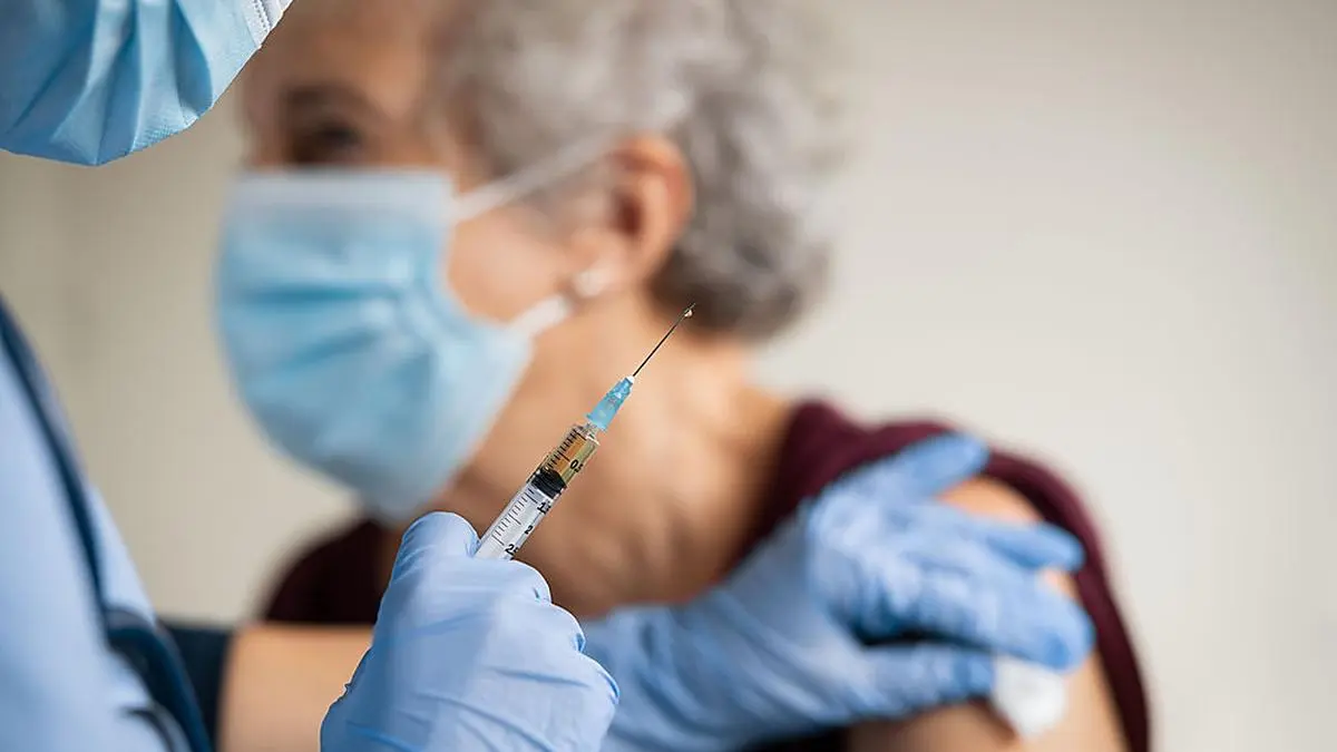 Close up of general practitioner hand holding vaccine injection while wearing face protective mask during covid-19 pandemic. Young woman nurse with surgical mask giving injection to senior woman at hospital. Close up of nurse holding syringe to vaccine old patient from covid19.