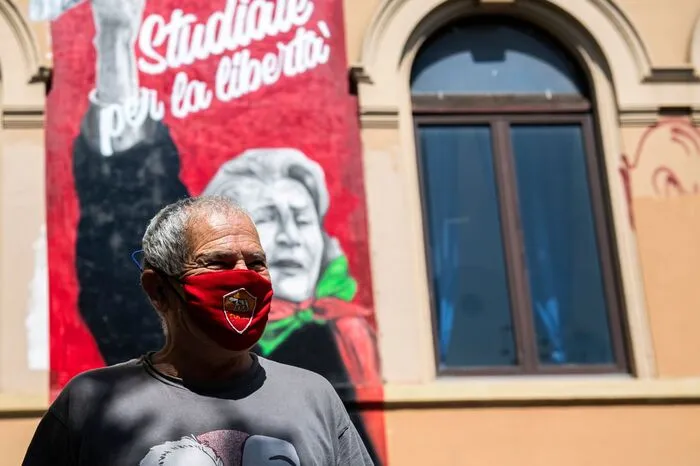 Ein maskierter Mann posiert neben einem Wandgemälde der verstorbenen Antifaschistin Tina Costa.  