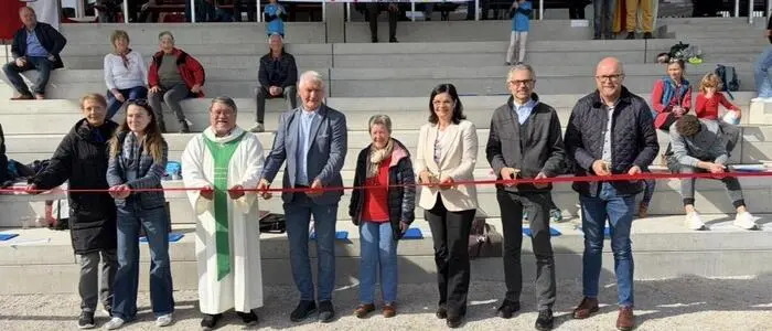 Mehrere Personen stehen vor einer Zuschauertribüne eines Sportplatzes und schneiden ein rotes Band durch