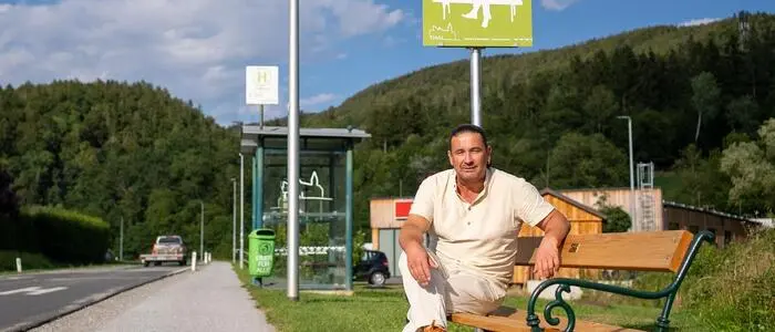 Er sitzt auf der Bank am Straßenrand und schaut in die Kamera, dahinter ein grünes Schild mit dem Titel „Mitfahrbankerl“. Im Hintergrund befindet sich eine Bushaltestelle.