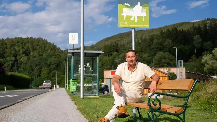 Er sitzt auf der Bank am Straßenrand und schaut in die Kamera, dahinter ein grünes Schild mit dem Titel „Mitfahrbankerl“. Im Hintergrund befindet sich eine Bushaltestelle.