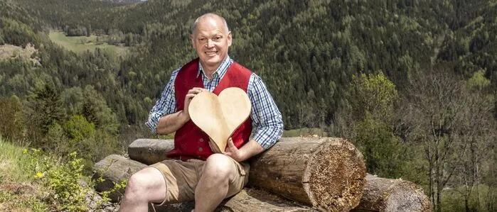 Ein Mann in Trachtengewand sitzt auf einem Holzstoß und hat ein Herz aus Holz in der Hand