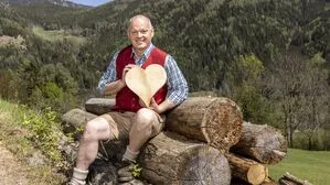 Ein Mann in Trachtengewand sitzt auf einem Holzstoß und hat ein Herz aus Holz in der Hand