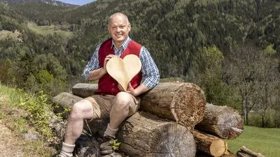Ein Mann in Trachtengewand sitzt auf einem Holzstoß und hat ein Herz aus Holz in der Hand
