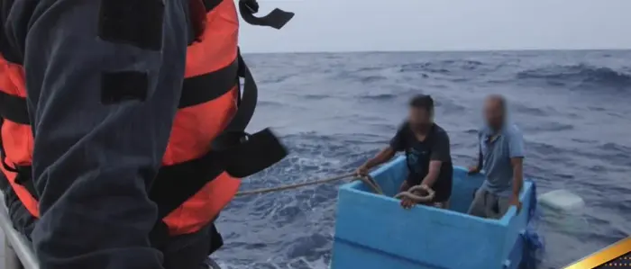 Zwei Männer in einer blauen Kühlbox treiben auf dem Meer, ein Mann mit Schwimmweste steht im Vordergrund