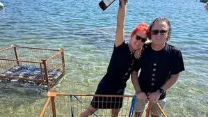 Eine Frau und ein Mann stehen im See hinter einem Metallgerüst, in dem Weinflaschen geschlichtet sind.