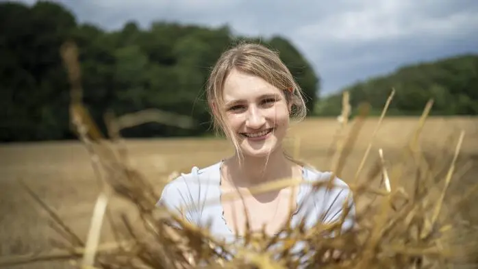 Eine junge Frau steht in einem Acker und hält Stroh in ihren Händen.