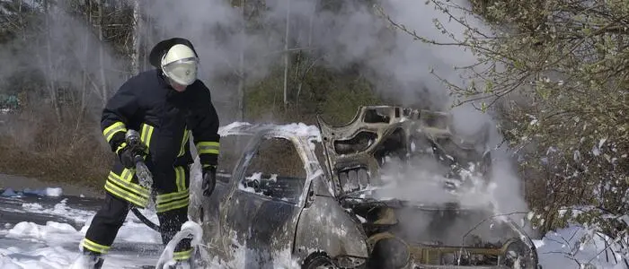 Ein Feuerwehrmann steht voll adjustiert auf einer Schicht Löschschaum, daneben steht ein rauchendes Autowrack