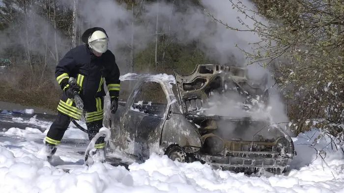 Ein Feuerwehrmann steht voll adjustiert auf einer Schicht Löschschaum, daneben steht ein rauchendes Autowrack