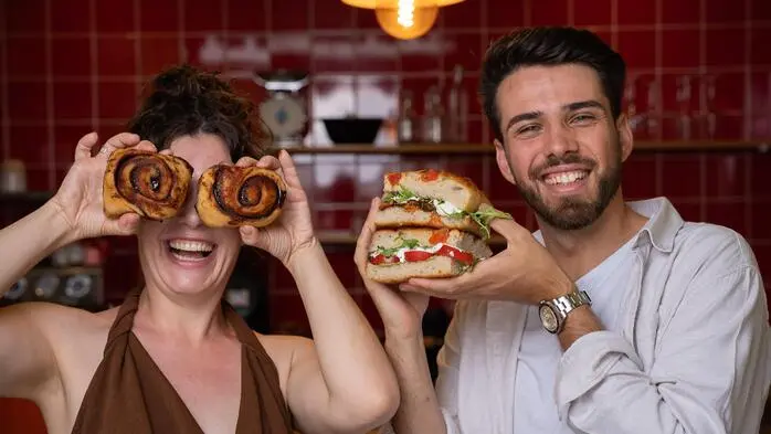 Die beiden gut gelaunten jungen Gastronomen in ihrem Lokal mit roten Fliesen im Hintergrund, sie hält Zimtschnecken vor ihre Augen, er präsentiert eine Focaccia
