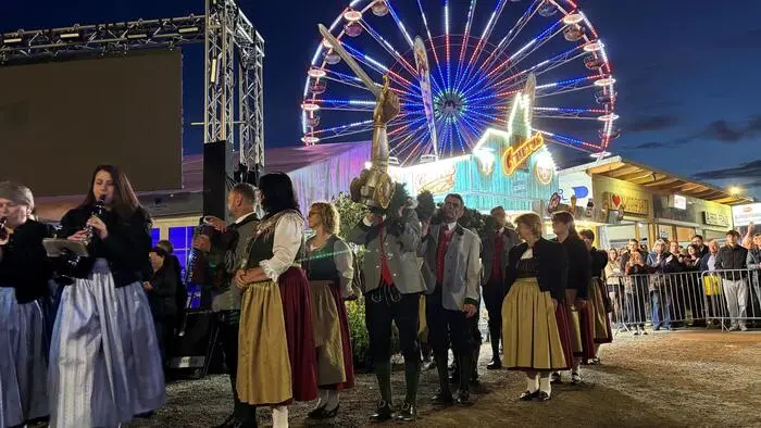 Männer in Tracht tragen einen langen Baumstamm und im Hintergrund ist ein Riesenrad