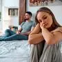Upset young woman in home clothes sitting on the bed, in the background sitting man. Family troubles.