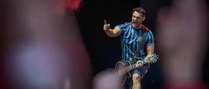 Paul Pizzera steht mit einem gestreiften Fußball-Trikot auf der Bühne. Er hat eine Gitarre umgehängt.