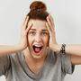 Young woman screaming in terror with hands on her head, mouth wide open looking in panic at the camera. Close up portrait of irritated female shouting, covering her ears, angry with noisy neighbours