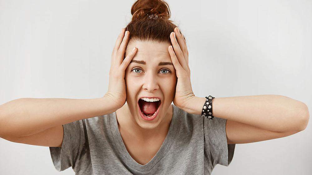 Young woman screaming in terror with hands on her head, mouth wide open looking in panic at the camera. Close up portrait of irritated female shouting, covering her ears, angry with noisy neighbours