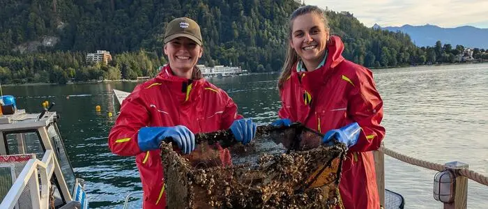 Zwei Frauen in roten Einsatzanzügen halten ein von Algen und Seetang bedecktes Netz in die Kamera. Sie stehen auf einem Steg, im Hintergrund sieht man einen See.